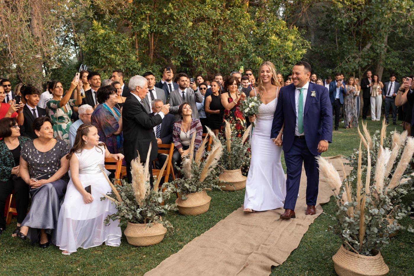 Bride and groom walk down a burlap aisle runner laid on the grass, past guests and baskets of pampas grass