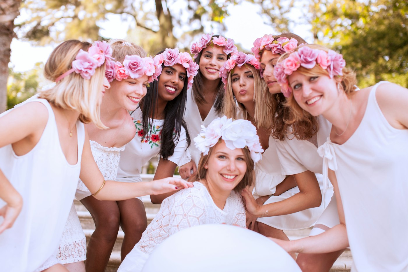 La future mariée coiffée d'une couronne blanche assise devant sept amies portant des couronnes de roses roses, toutes souriantes en plein air