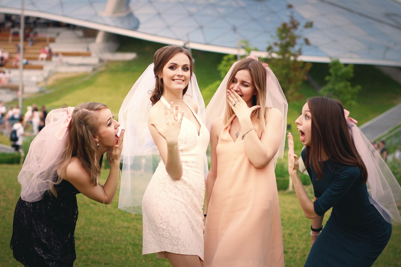 La future mariée montre sa bague de fiançailles, trois amies coiffées de petits voiles font mine d'être surprises dans un parc