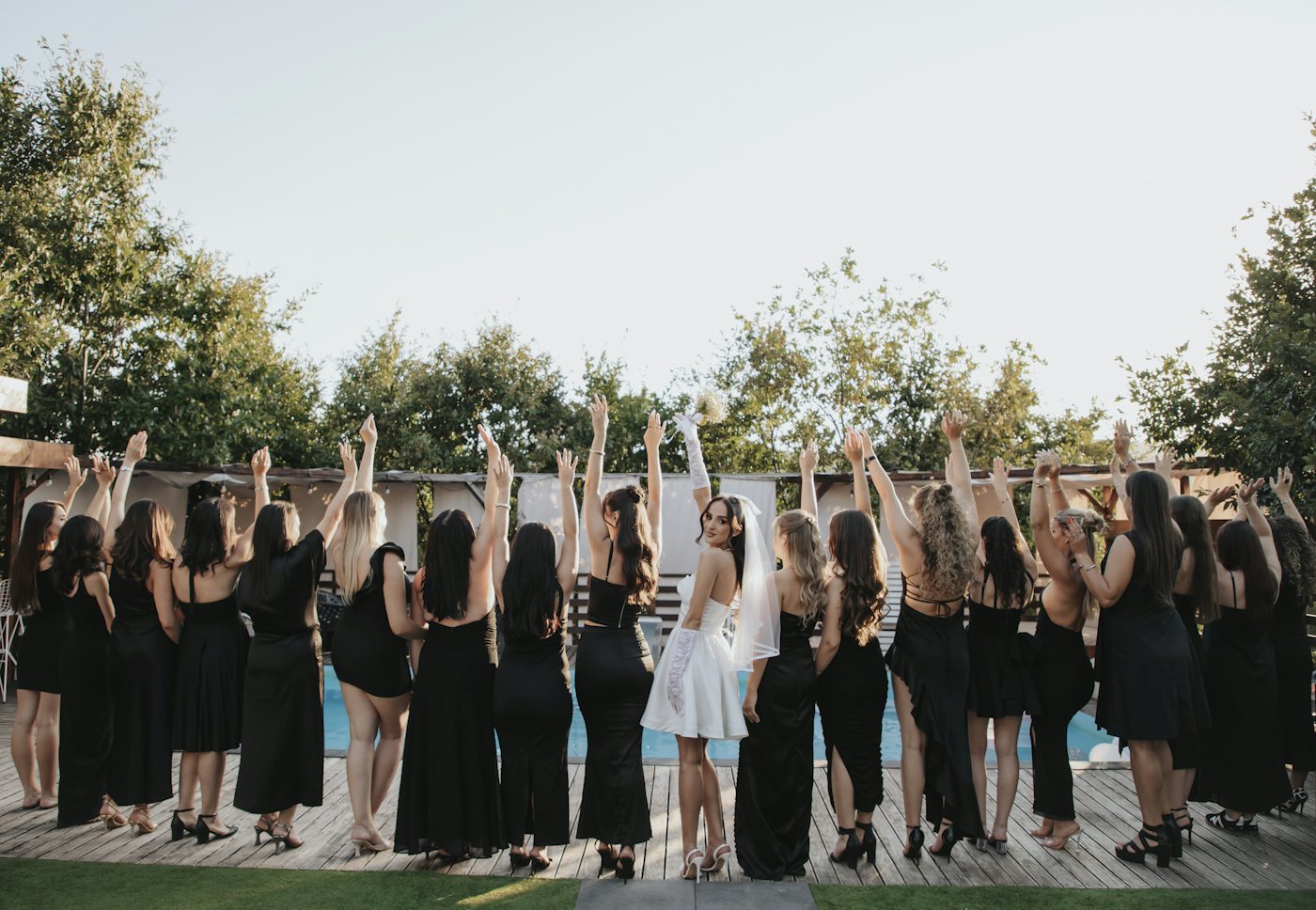 La future mariée en robe blanche courte et voile lève les bras avec ses amies en robes noires au bord d'une piscine