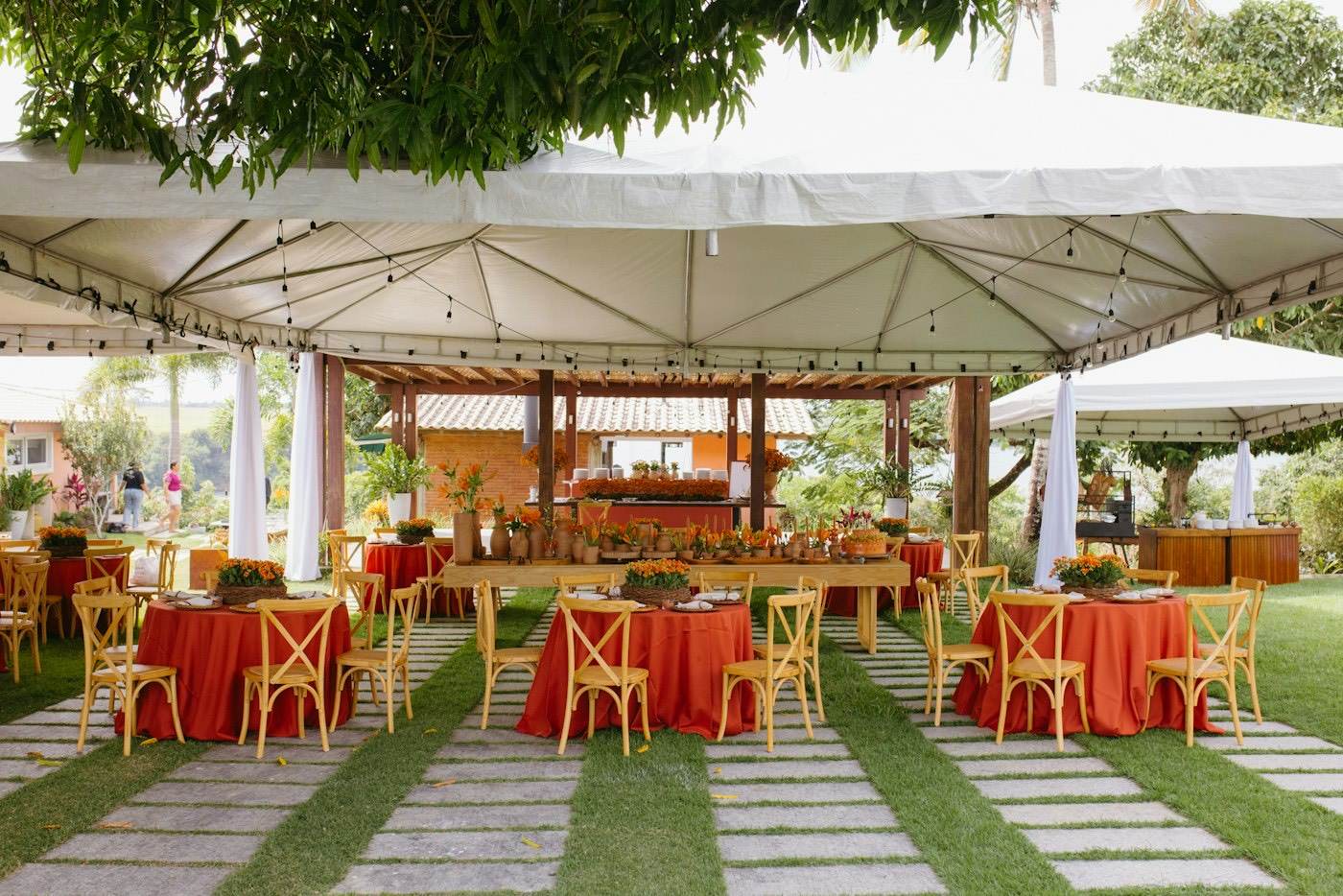 Une tente blanche de location sur la pelouse avec des tables rondes en nappes orange et des chaises en bois pour une réception de mariage au jardin
