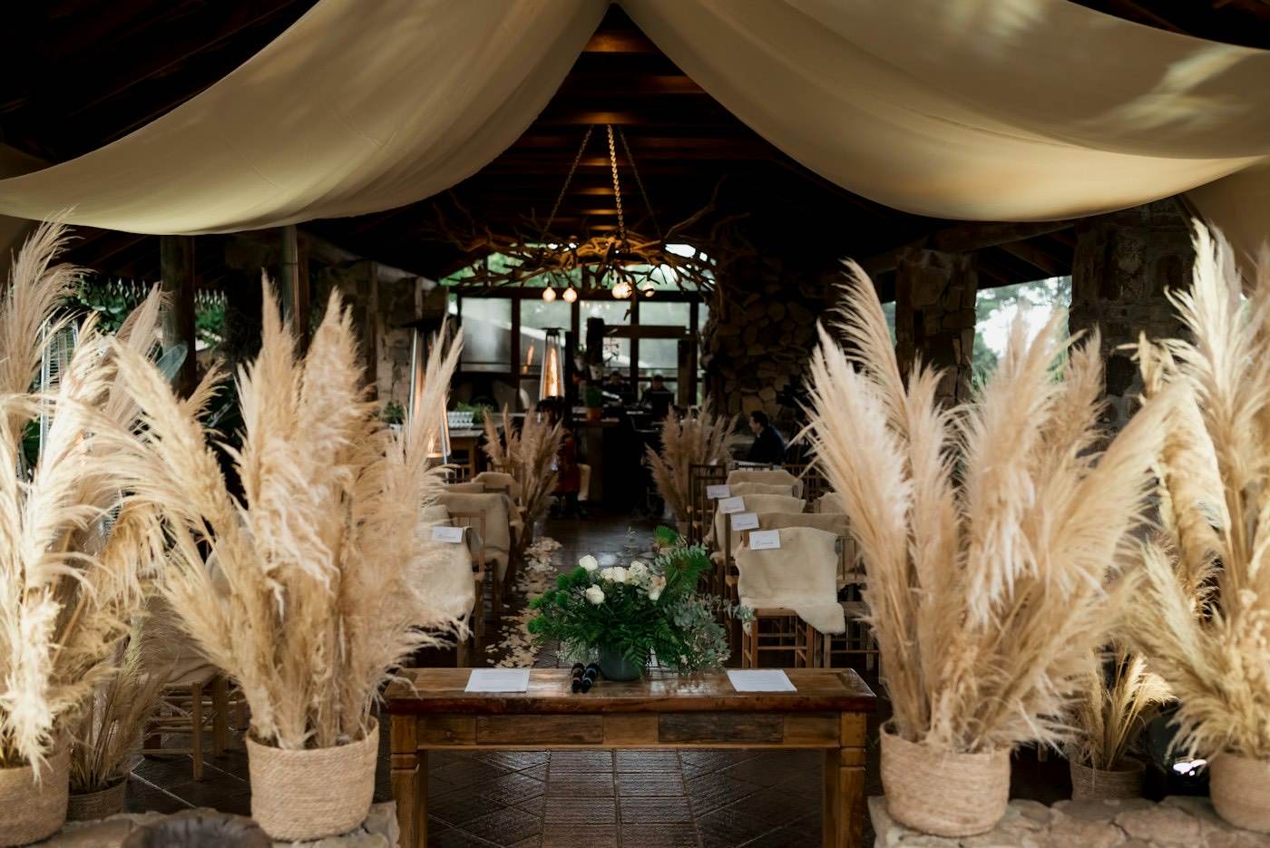 A ceremony aisle lined with tall pampas grass in woven baskets under cream fabric drapes