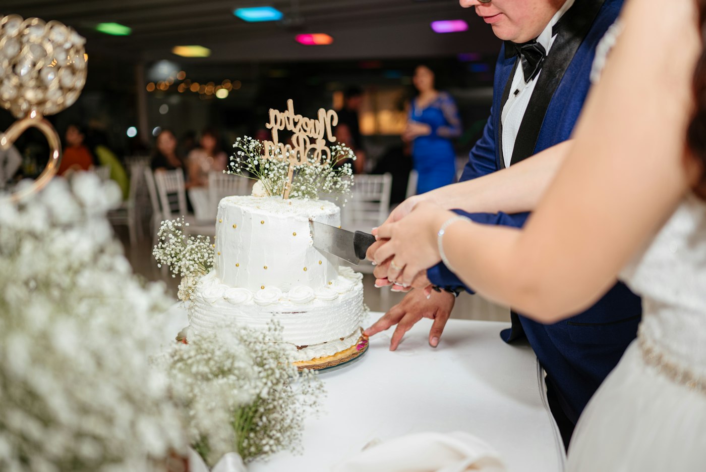 A bride and groom in a blue tuxedo cut a small two-tier white buttercream cake topped with baby's breath at an evening reception