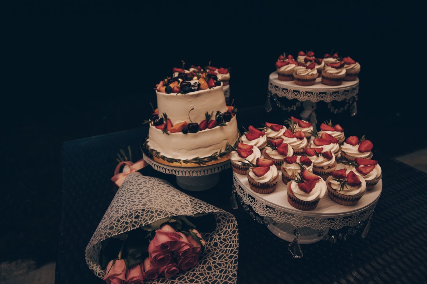 A small two-tier buttercream cake topped with berries next to two stands of strawberry cupcakes and a wrapped bouquet of pink roses