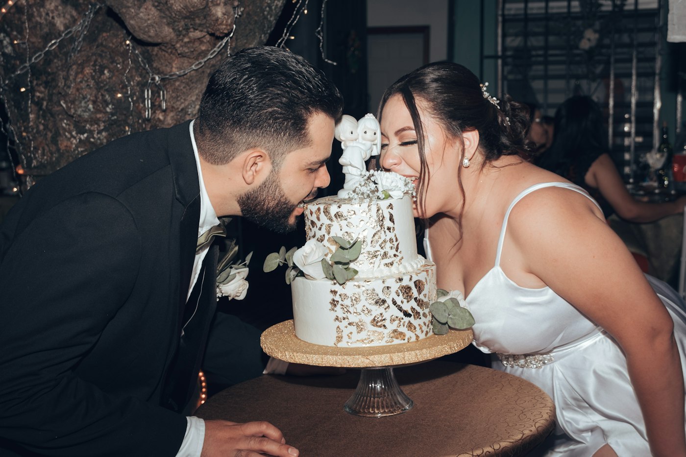 A laughing bride and groom lean in to take a bite from either side of a small two-tier white and gold cake