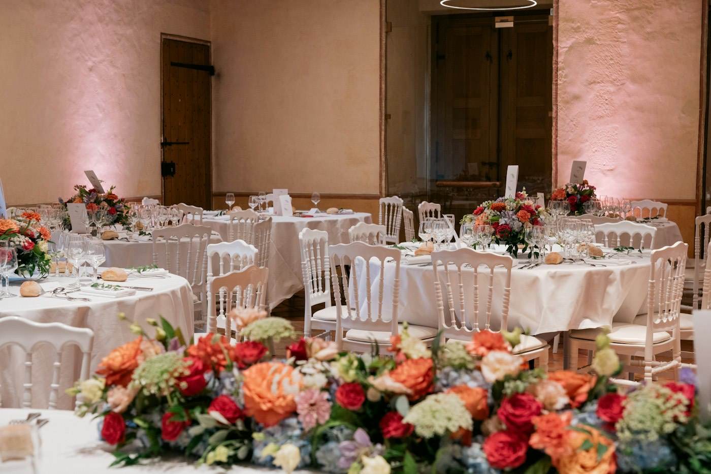 Des tables rondes de réception de mariage avec de grandes compositions de fleuriste en roses et hortensias orange, rouges et bleus