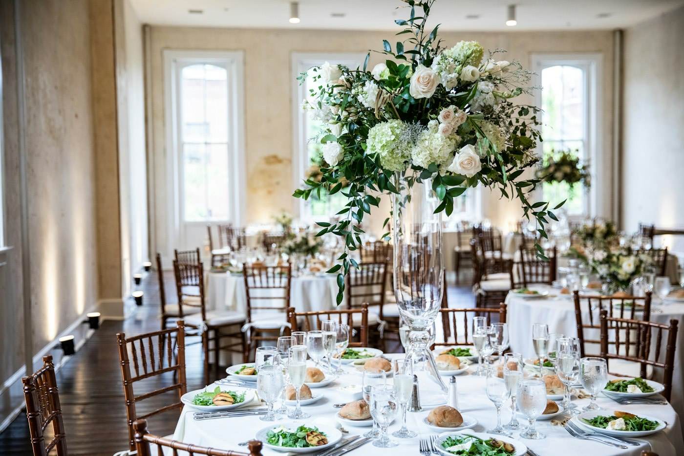 Un grand centre de table de fleurs blanches sur un pied en verre, avec des compositions basses sur les tables derrière