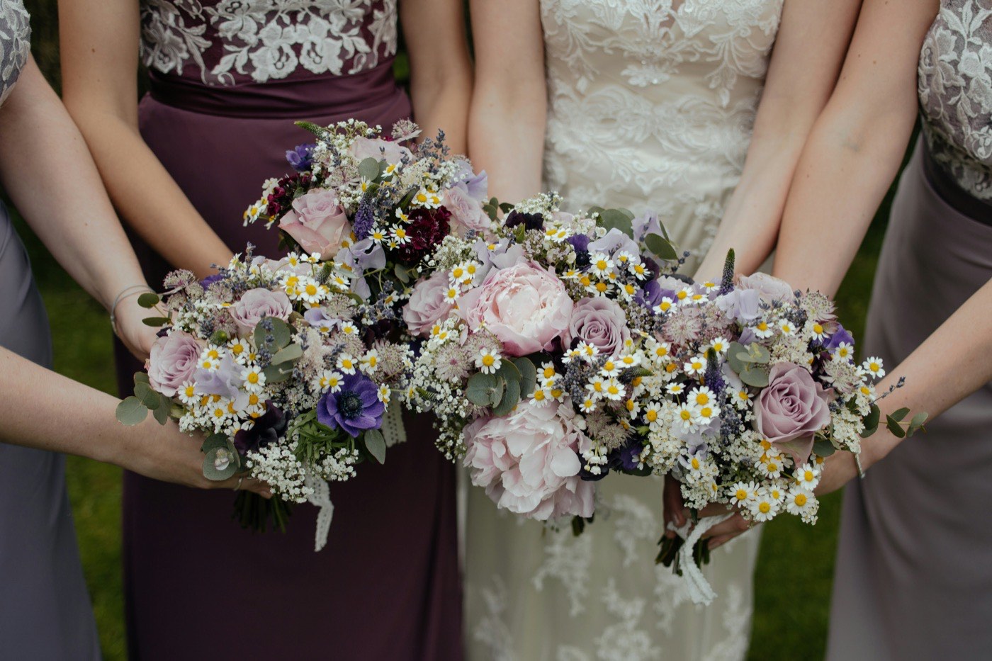 Quatre demoiselles d'honneur en mauve et gris et la mariée en dentelle rapprochent leurs bouquets roses, lilas et blancs