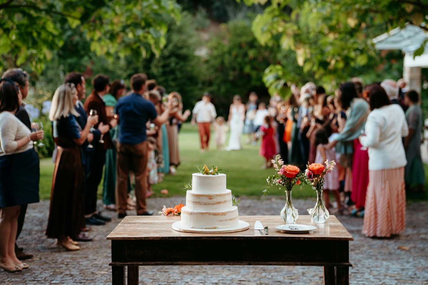 Un gâteau à trois étages sur une table en bois à côté de deux petits vases de roses orange, avec les invités sur la pelouse derrière