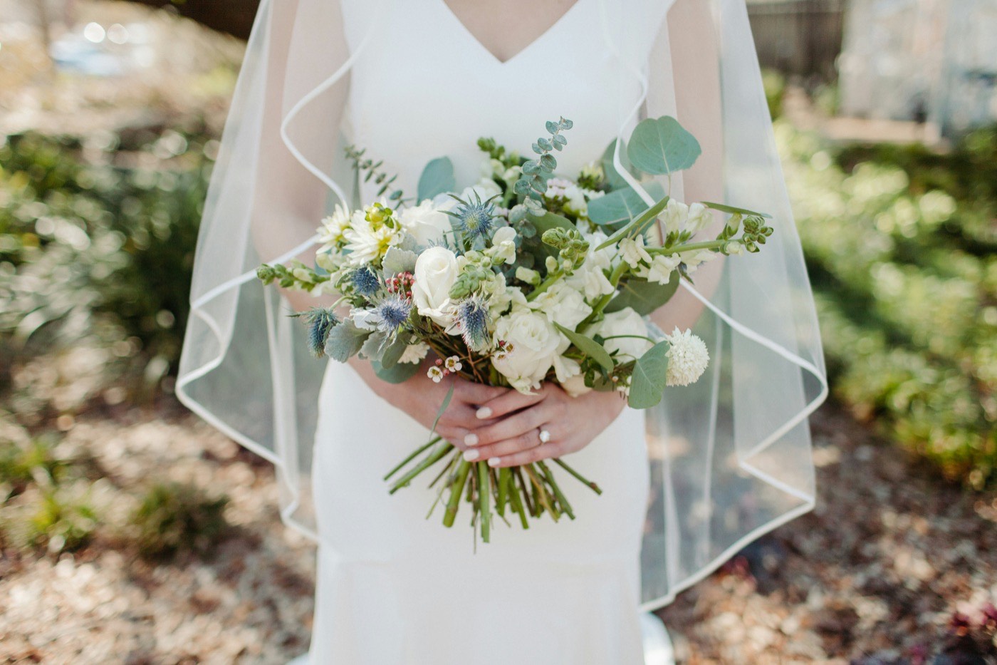 Une mariée en robe blanche et voile tient un bouquet souple de roses blanches, de chardons et d'eucalyptus dans un jardin