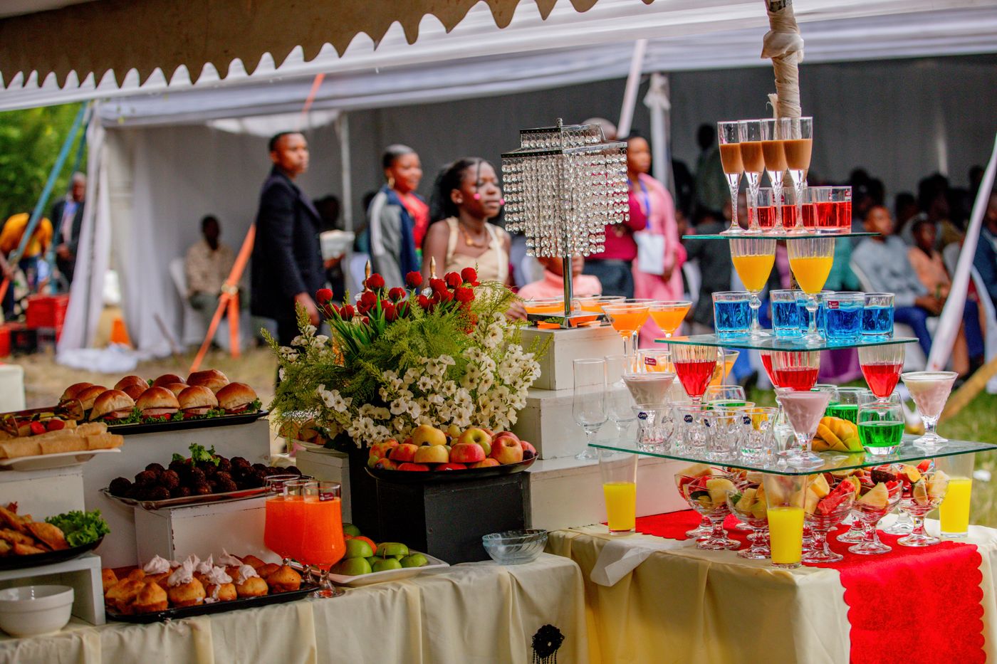 Buffet de mariage sous chapiteau avec mini burgers, fruits et verres colorés, des invités discutent en arrière-plan