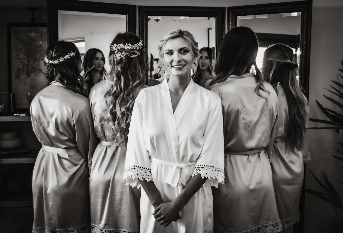 Une mariée en peignoir blanc devant un mur de miroirs, entourée de ses amies avec des fleurs dans leurs cheveux ondulés