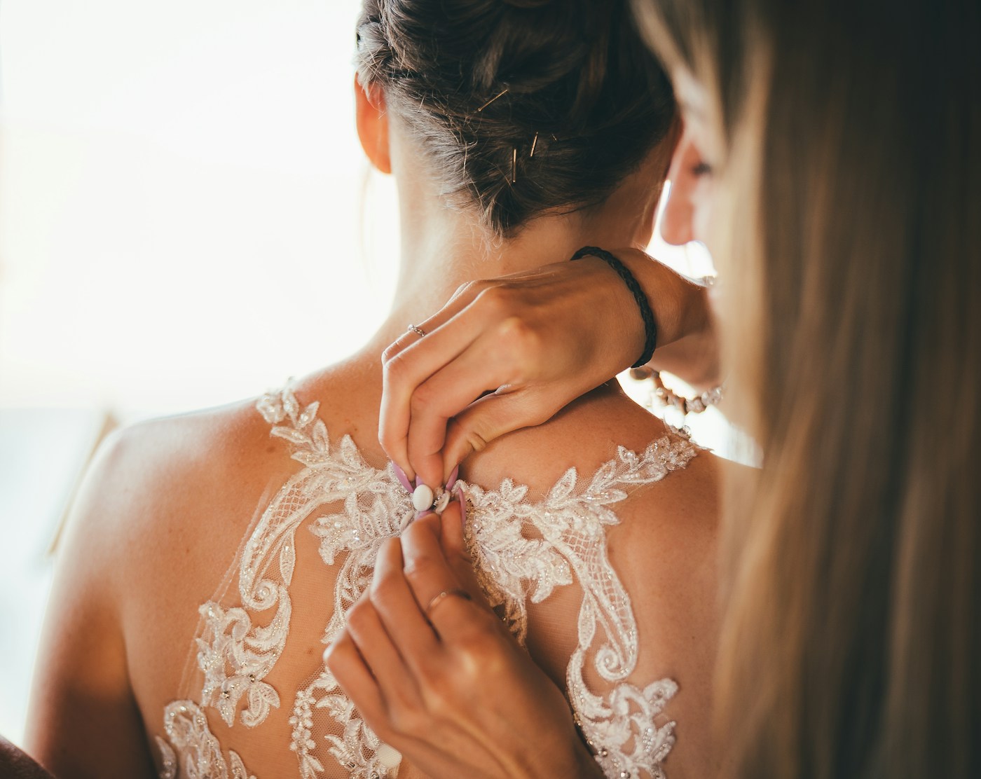 Une amie ferme les boutons du dos en dentelle de la robe de la mariée, coiffée d'un chignon simple tenu par des épingles