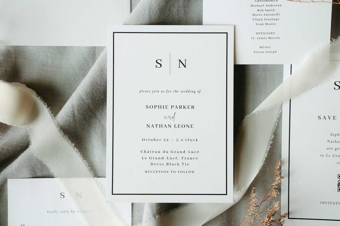Un faire-part blanc non plié, avec un fin cadre noir et une typographie sobre, posé sur du lin gris avec un ruban de soie effiloché et des fleurs séchées