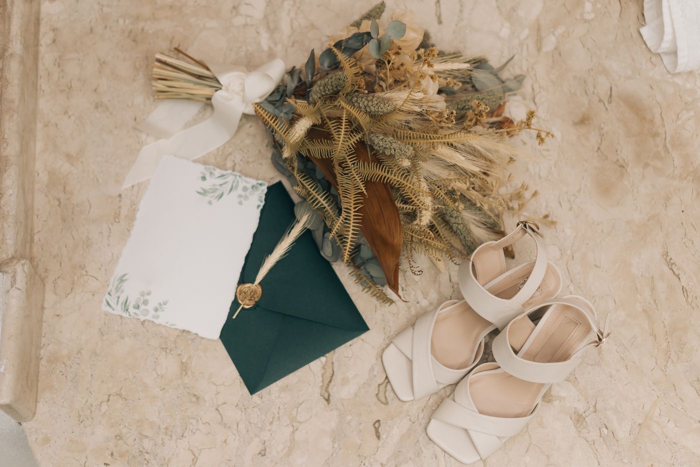 Une enveloppe vert sapin fermée par un cachet de cire doré et un épi séché, à côté d'un bouquet de fleurs séchées et de sandales de mariée crème sur du marbre