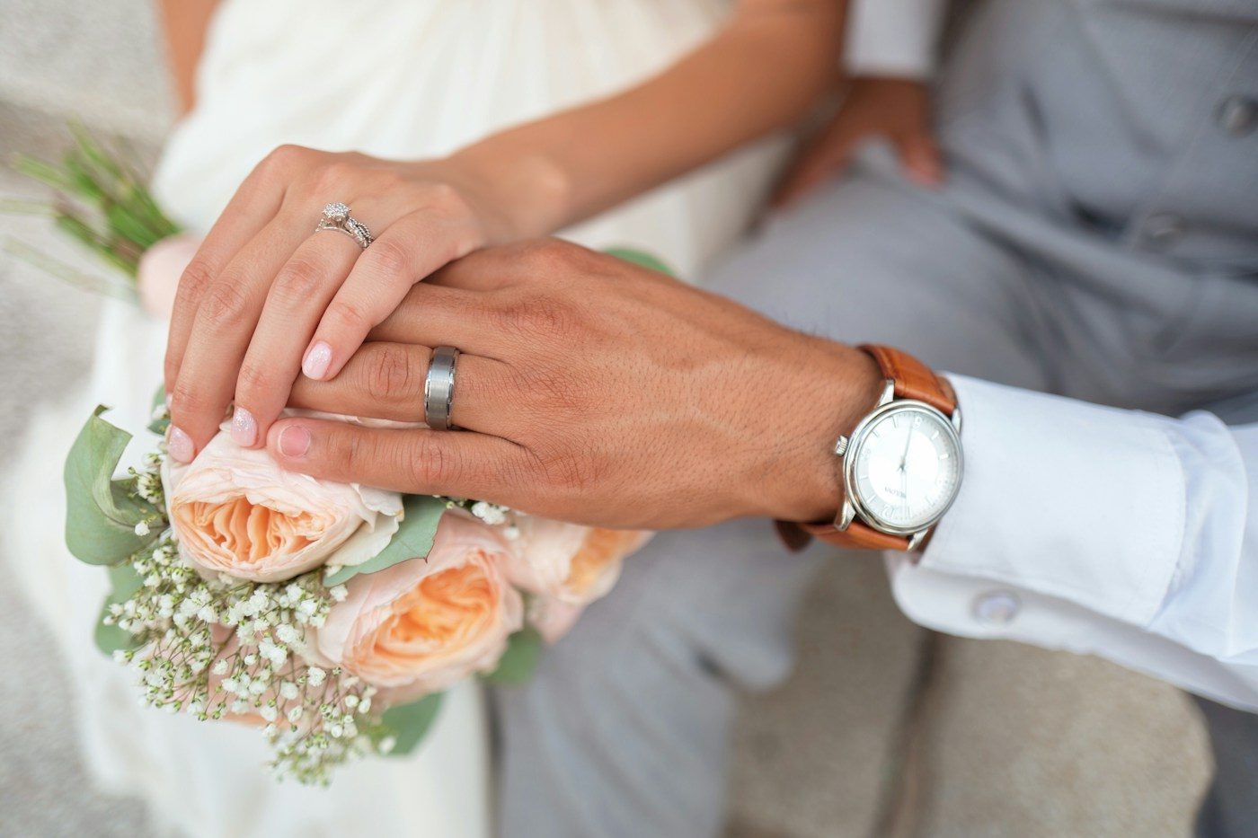 Die Braut legt ihre Hand mit Verlobungsring auf die Hand des Bräutigams, der einen gebürsteten grauen Ring trägt, darunter ein Strauß mit apricotfarbenen Rosen