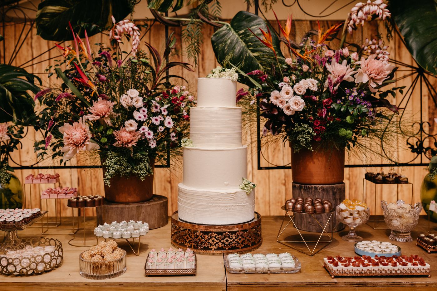A tall white wedding cake on a wooden dessert table with large flower arrangements and small trays of sweets around it