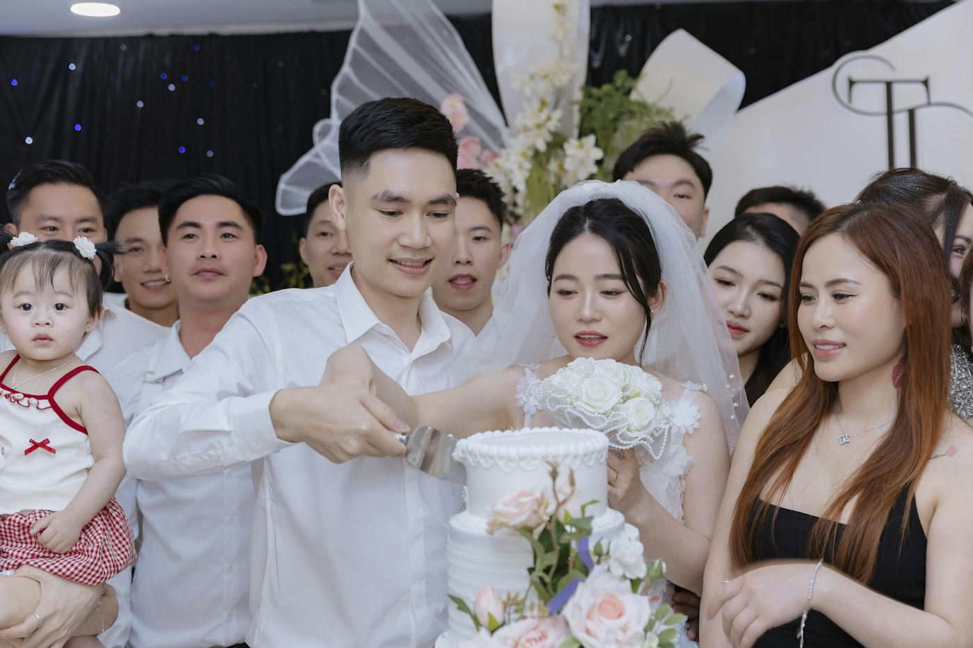 A bride and groom cut a white two-tier cake with pink roses while family and friends crowd in to watch