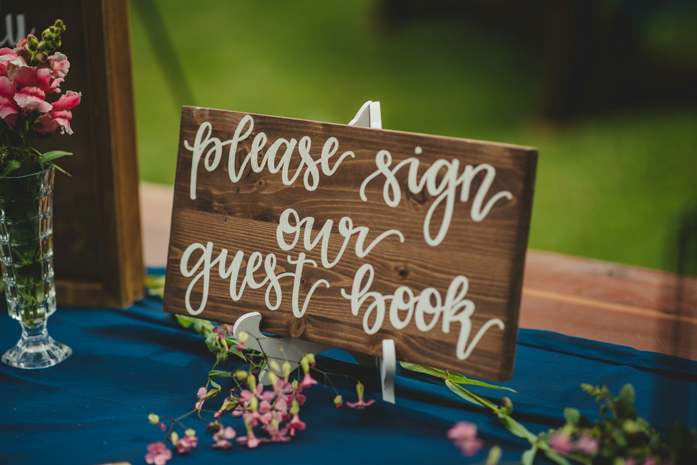 A hand-lettered wooden sign asking guests to sign the guest book stands on a table with a blue cloth and pink flowers