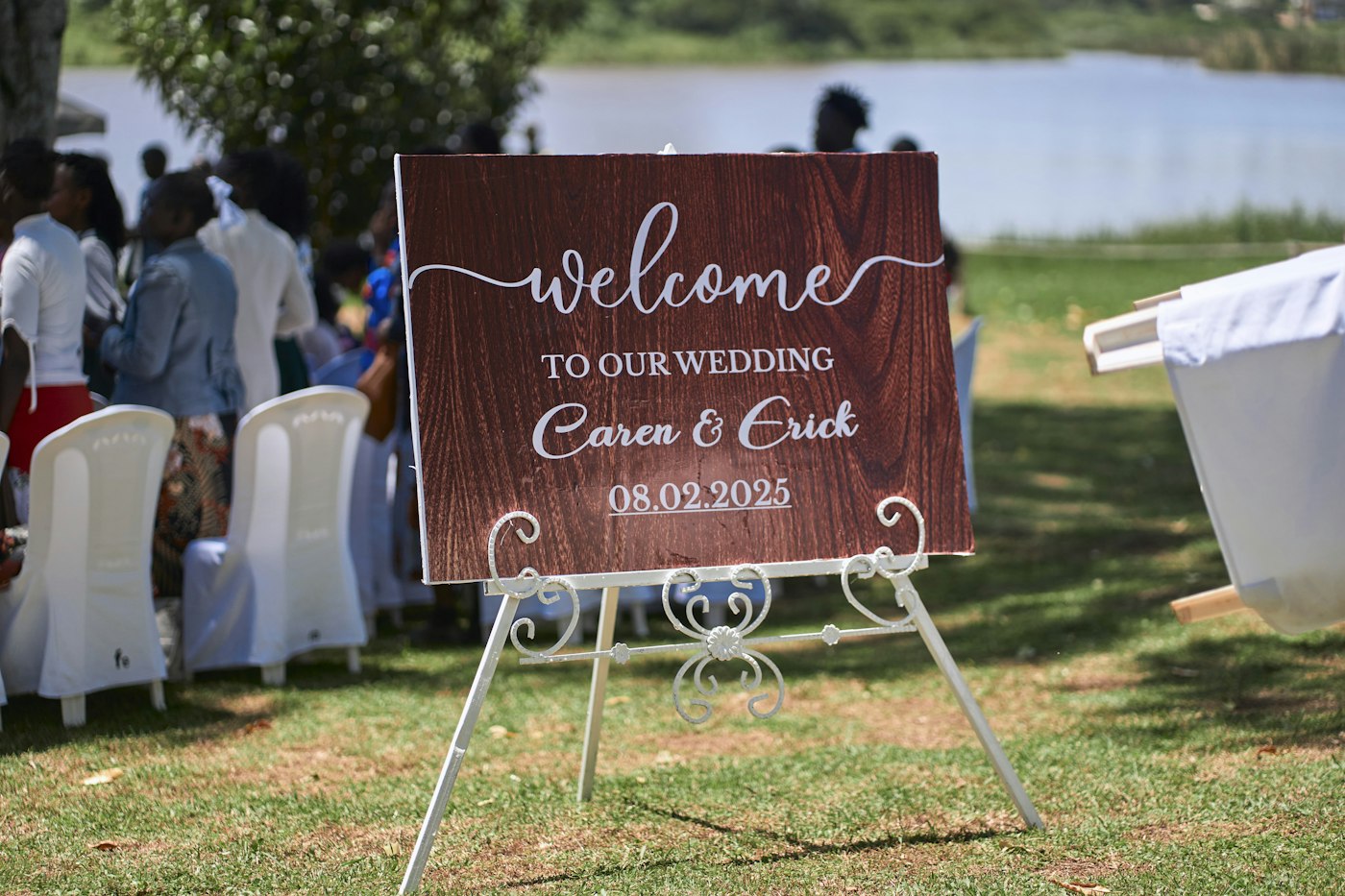A wood-print welcome sign with the couple's names and date stands on a white easel on the lawn while guests gather by the lake