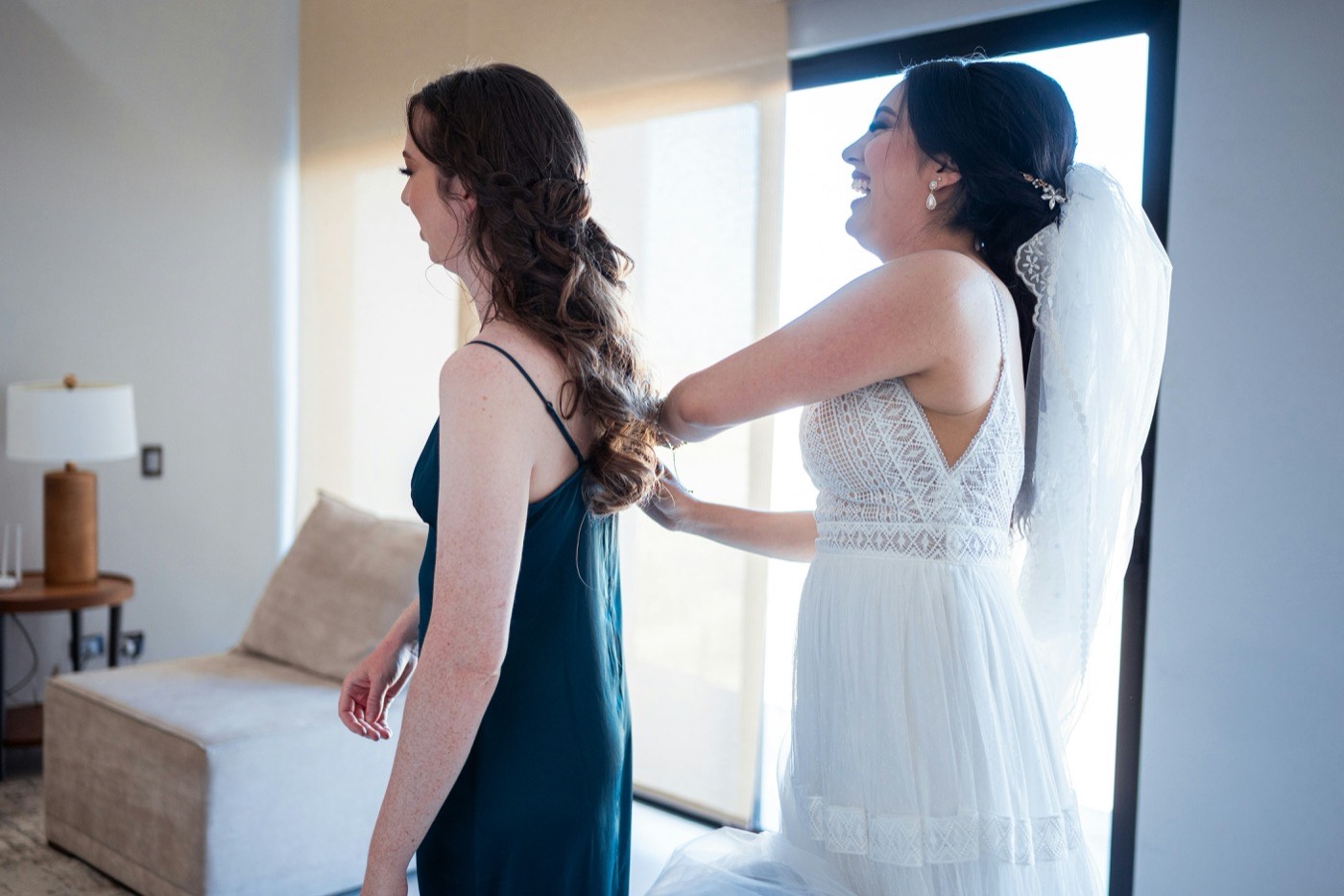 The bride, already in her dress and veil, laughs as she fixes a bridesmaid's hair by a sunny window