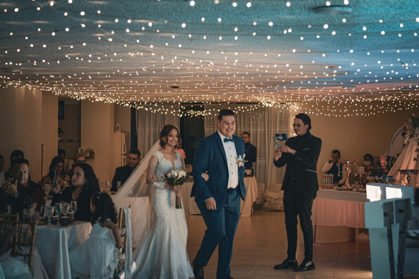 The bride and groom walk arm in arm into their reception under a ceiling of fairy lights while guests watch from the tables