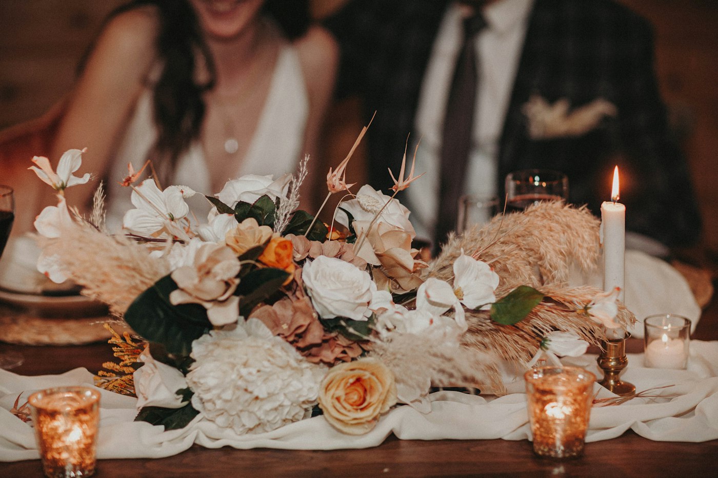 A bride and groom behind a dried flower centerpiece with small glass votives and a white taper candle in a brass holder