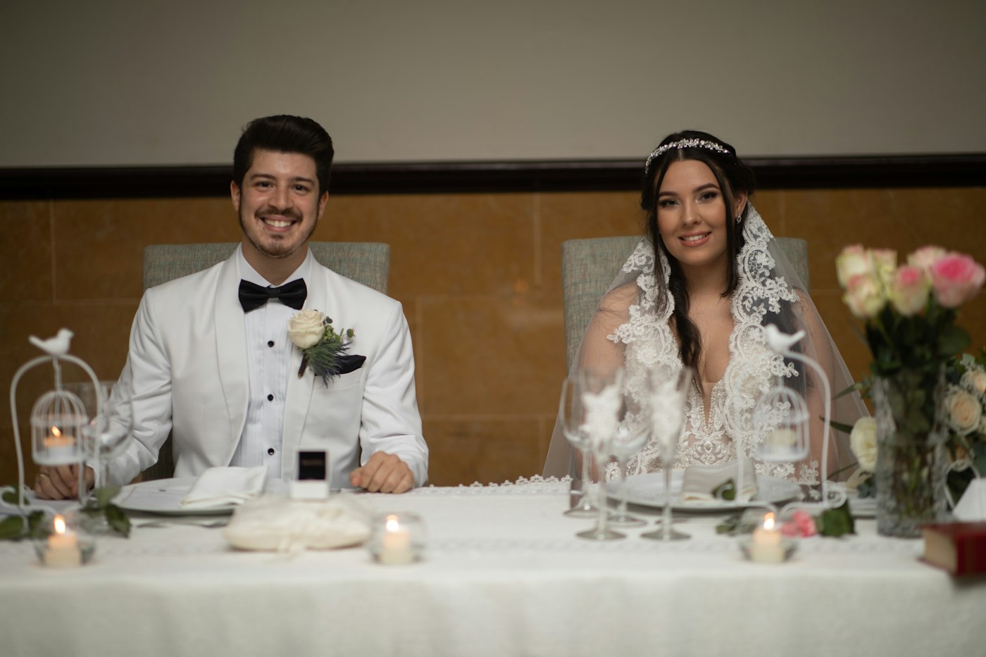 A smiling bride and groom at their head table with small glass tealight holders, white birdcage lanterns and a glass vase of pink roses