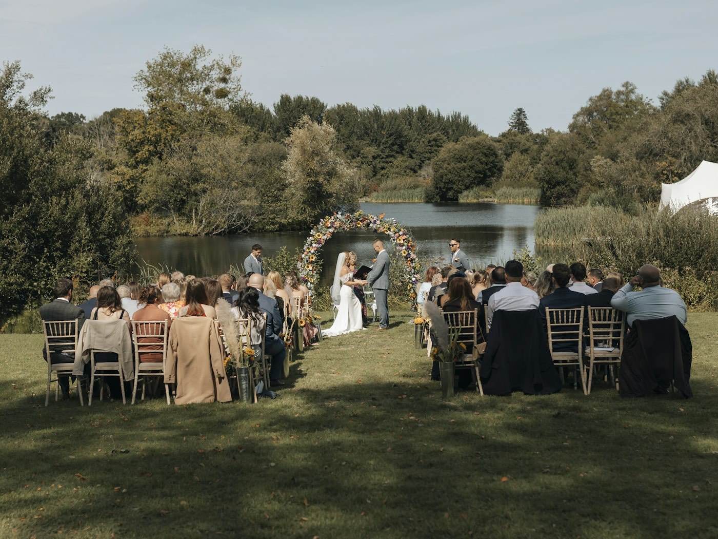 Invitados sentados en el césped asisten a una ceremonia al aire libre bajo un arco de flores junto a un lago a comienzos del otoño