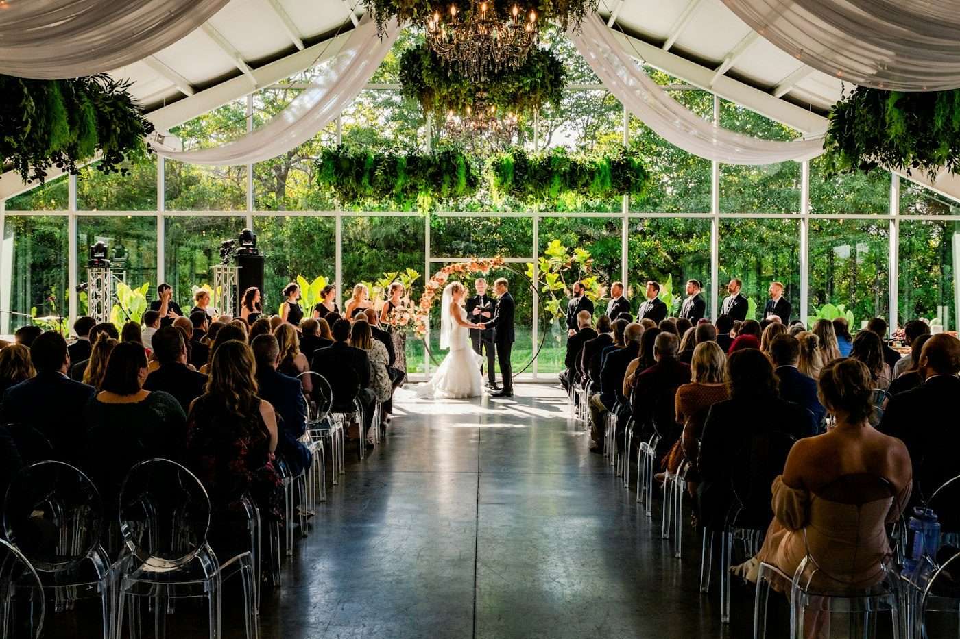 Guests on clear chairs watch a couple marry in a glass-walled garden pavilion hung with greenery and white drapes