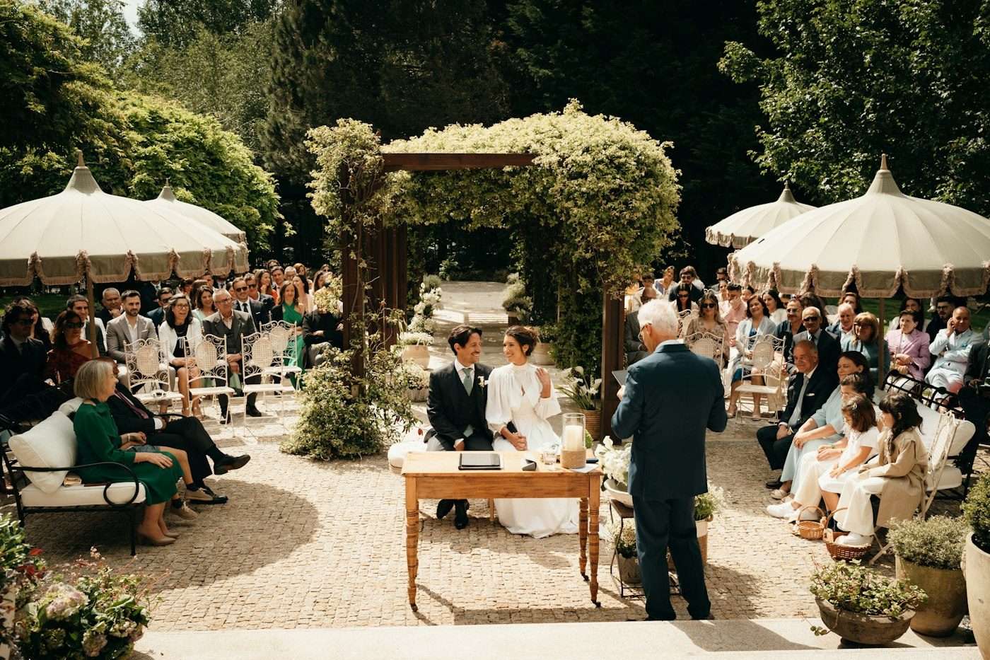 A couple sits at a small wooden table under a leafy arch while guests watch from garden chairs and big cream parasols