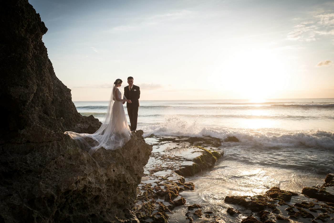 A bride with a long veil and a groom in a dark suit hold hands on a rock above the ocean at sunset