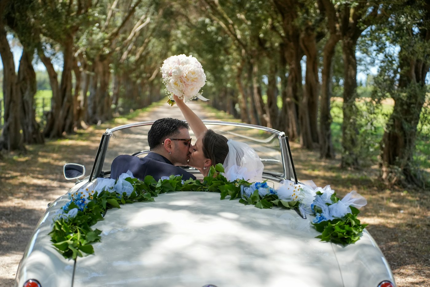 A bride raises her peony bouquet while kissing the groom in a convertible decorated with greenery on a tree-lined road