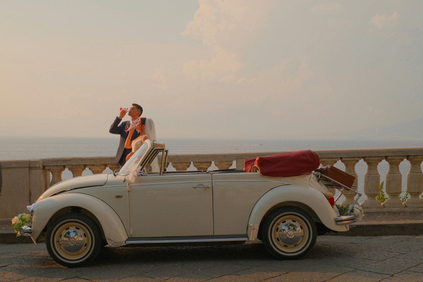A bride and groom raise a glass beside a vintage convertible with a suitcase strapped to the back, the sea behind them