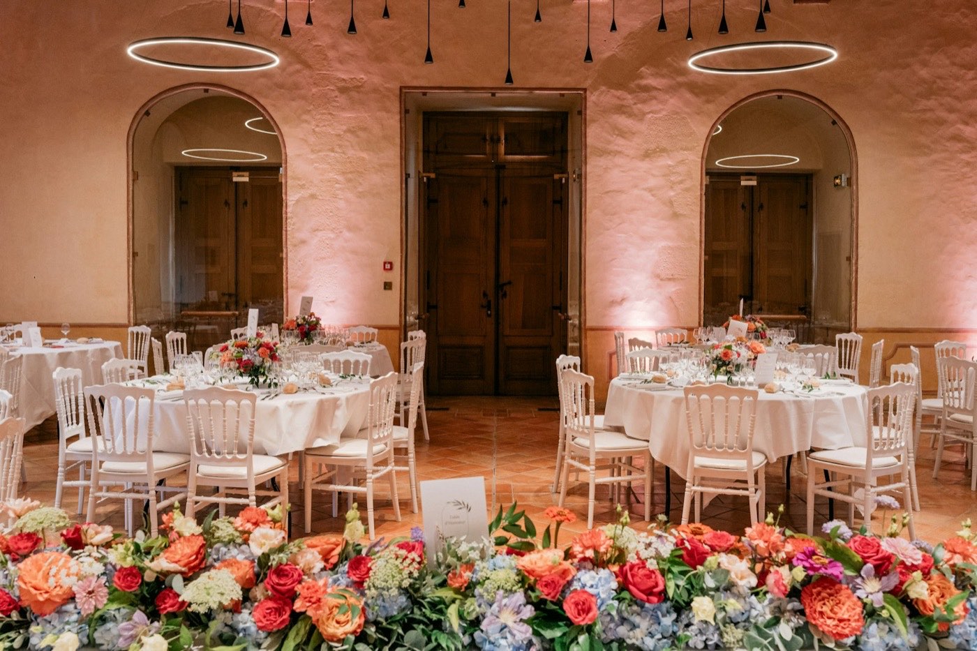 Round reception tables each with one flower centerpiece, behind a long low row of red, orange and blue flowers
