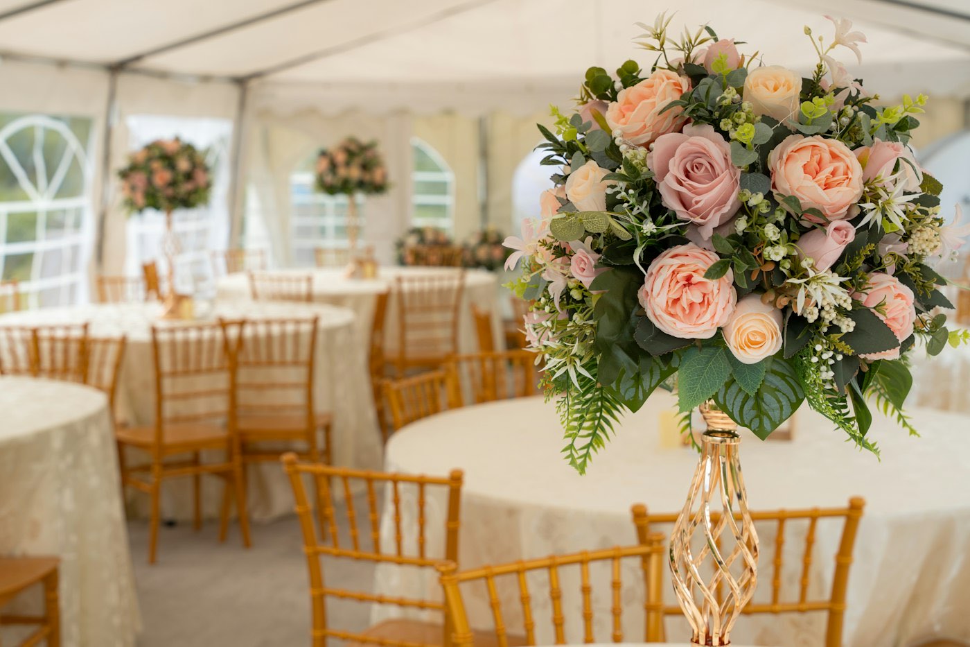 A tall arrangement of peach roses on a gold stand on a round table in a white wedding tent, more tables behind
