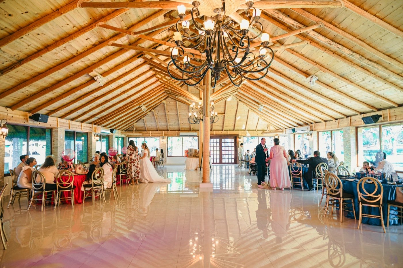 Wedding guests seated at round tables with flower centerpieces in a wooden hall with a chandelier, the bride in white among them