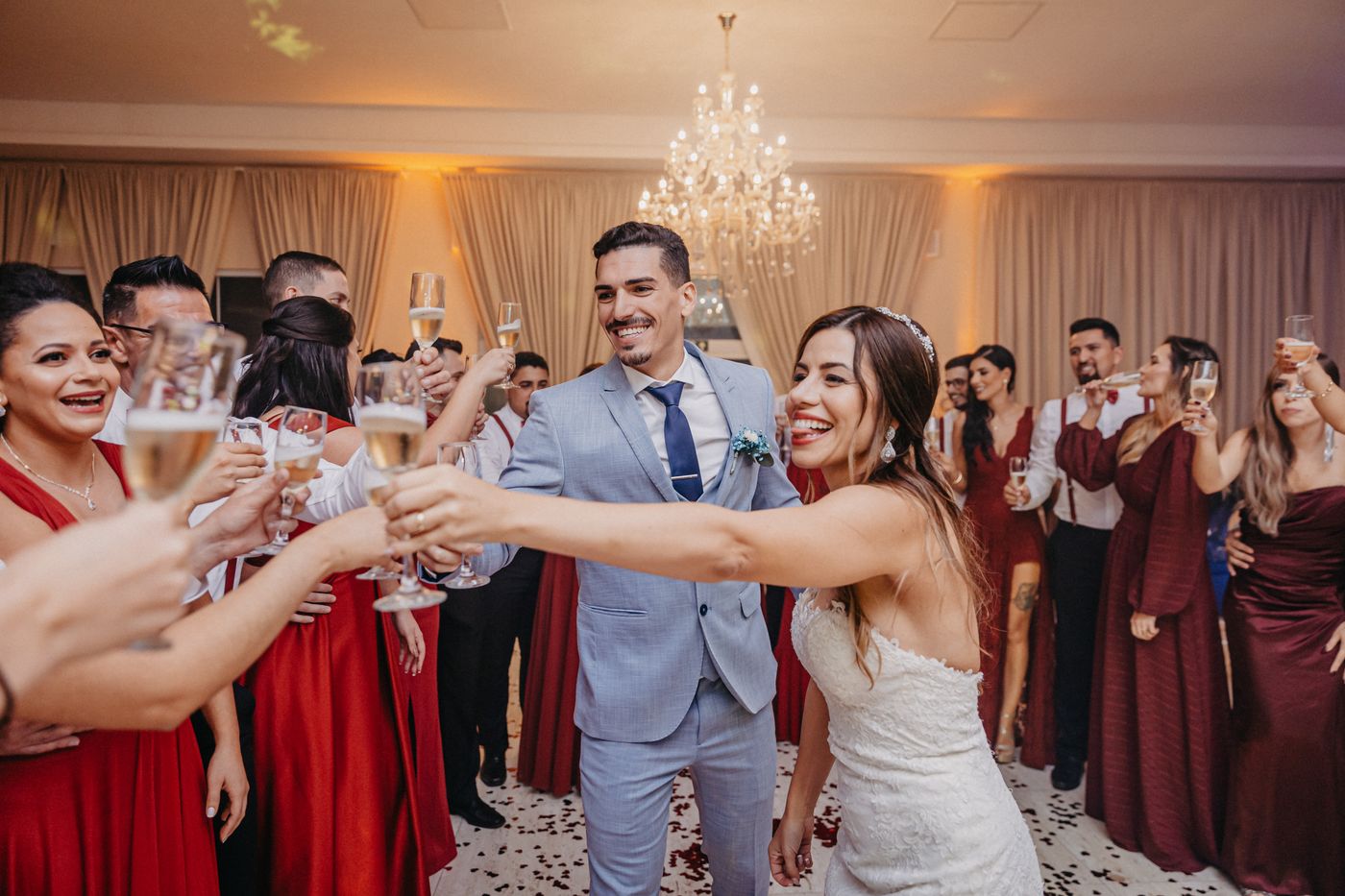 Los novios brindan con cava entre risas junto a damas de honor vestidas de rojo e invitados en un salón con lámpara de araña
