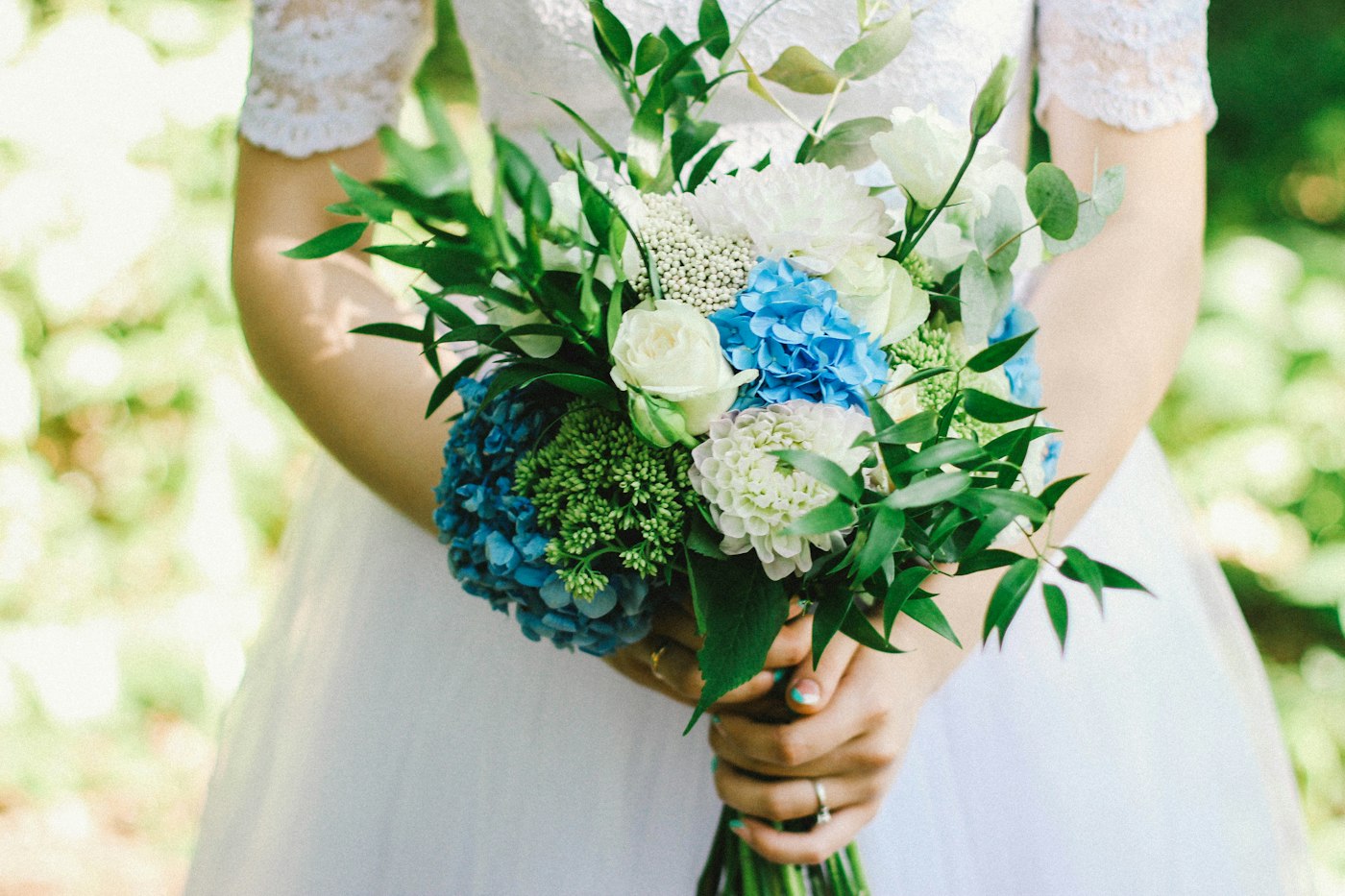 Eine Braut im Spitzenkleid hält einen locker gebundenen Strauß aus blauer Hortensie, weißen Rosen, Dahlien und langen grünen Blättern