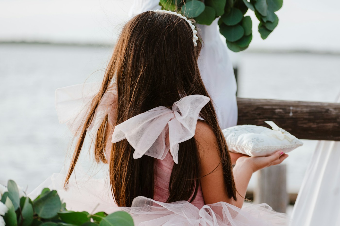 A flower girl in a pink tulle dress holds a ring pillow by the water at an outdoor ceremony