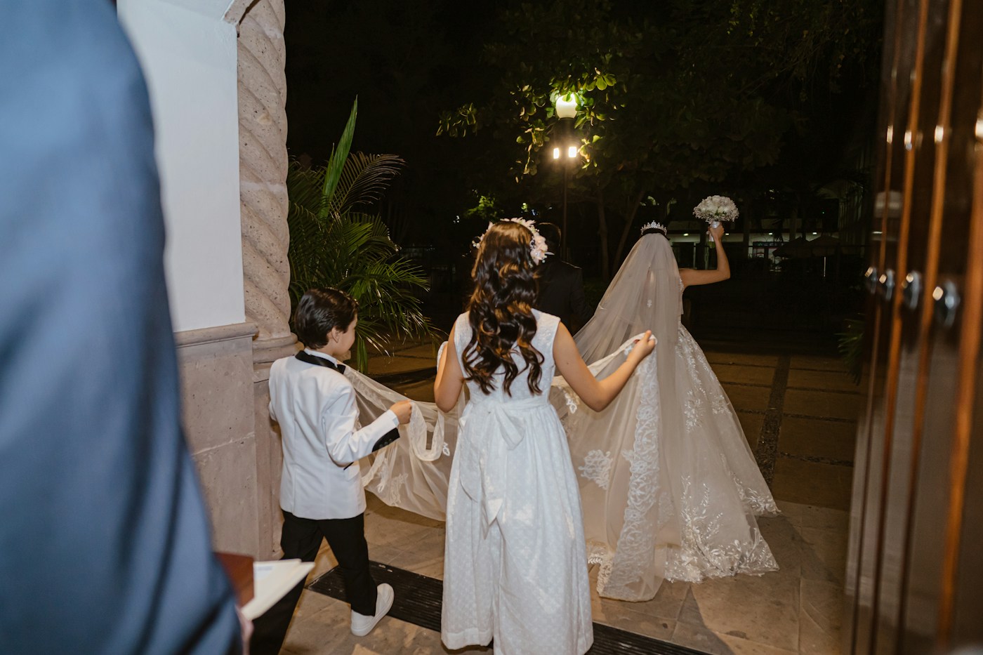 A young ring bearer and a flower girl carry the bride's long veil as she walks out into the evening with her bouquet raised