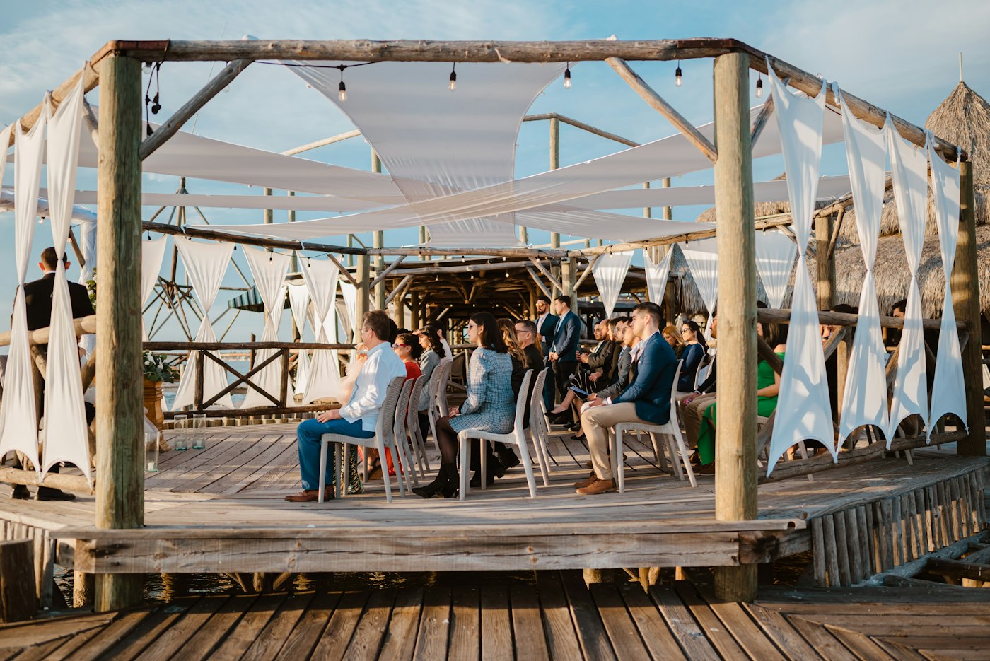 Guests in smart clothes sit on white chairs on a wooden deck by the water, under a simple frame draped with white fabric