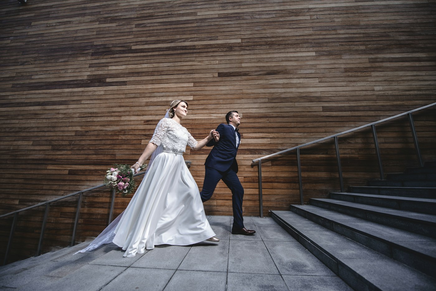 A bride in a lace top and satin skirt and a groom in a navy suit walk hand in hand past a modern wooden wall and stone steps