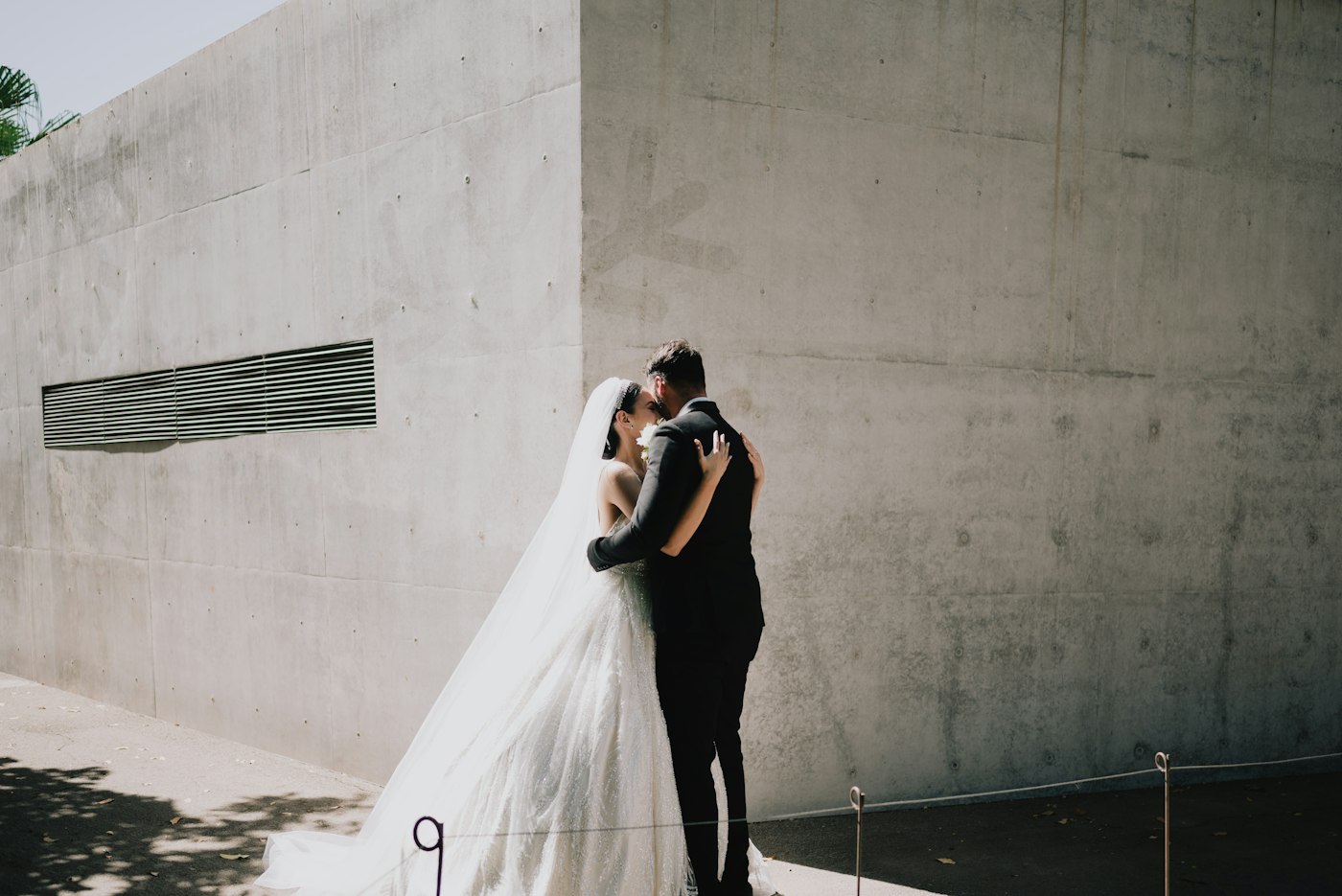 A bride in a long veil and a groom in a black suit embrace in front of a plain concrete wall in bright sunlight
