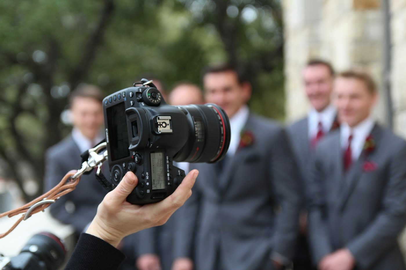 A photographer holds up a camera in front of a row of smiling groomsmen in grey suits with red boutonnieres