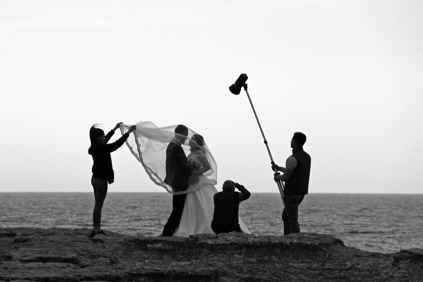 Black and white photo of a wedding shoot on a sea cliff: an assistant lifts the bride's veil, a photographer kneels and another holds a light on a pole