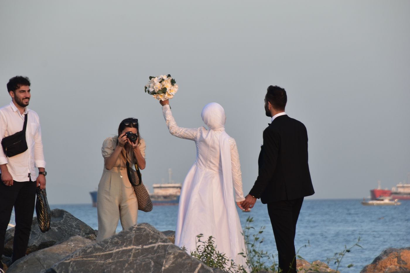 A photographer crouches on the rocks by the sea to photograph a bride raising her bouquet while holding the groom's hand