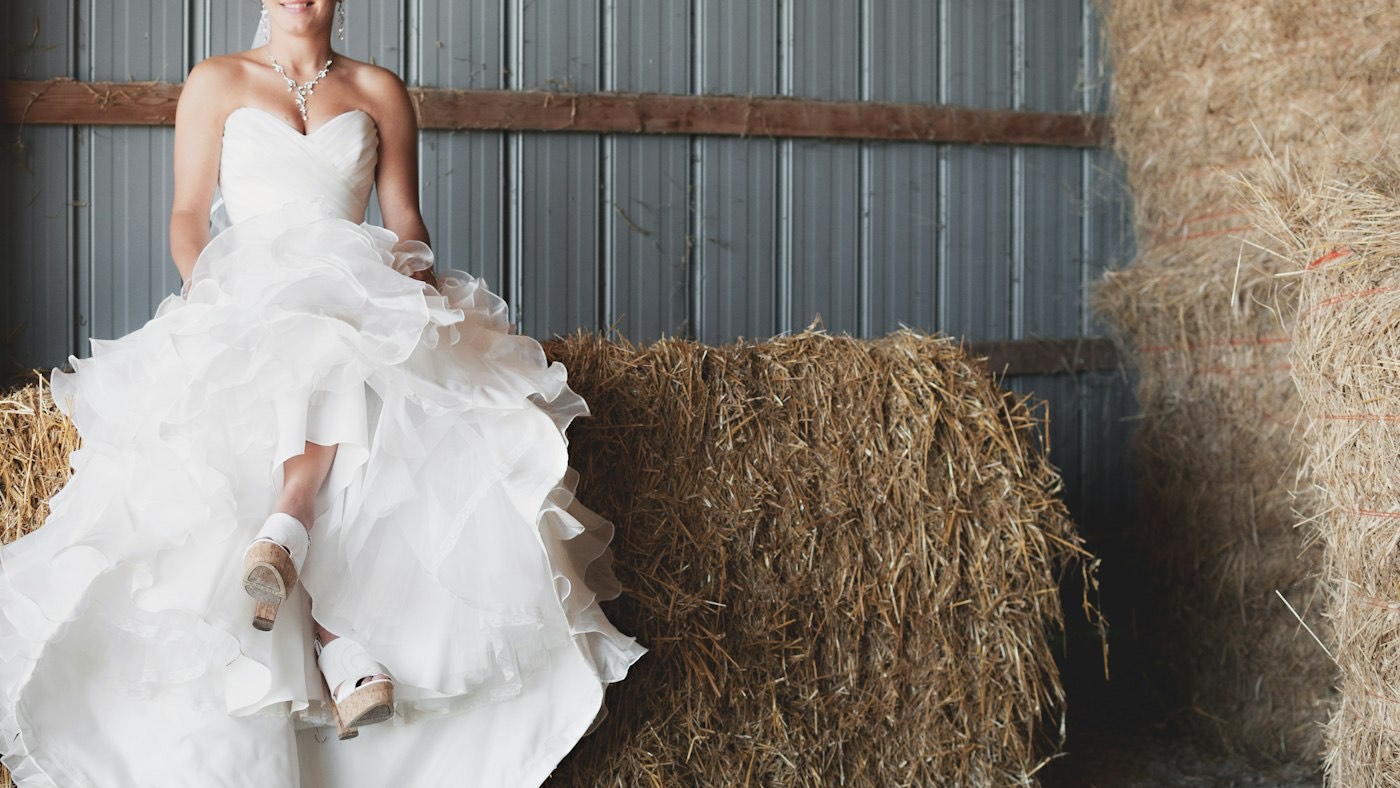 A bride in a ruffled wedding gown sits on a hay bale in a barn with corrugated metal walls