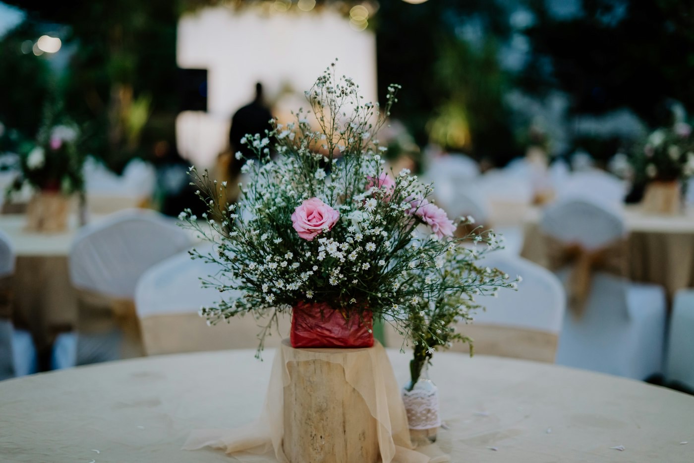 A simple centerpiece of baby’s breath and pink roses on a log slice at a reception table with sashed chairs