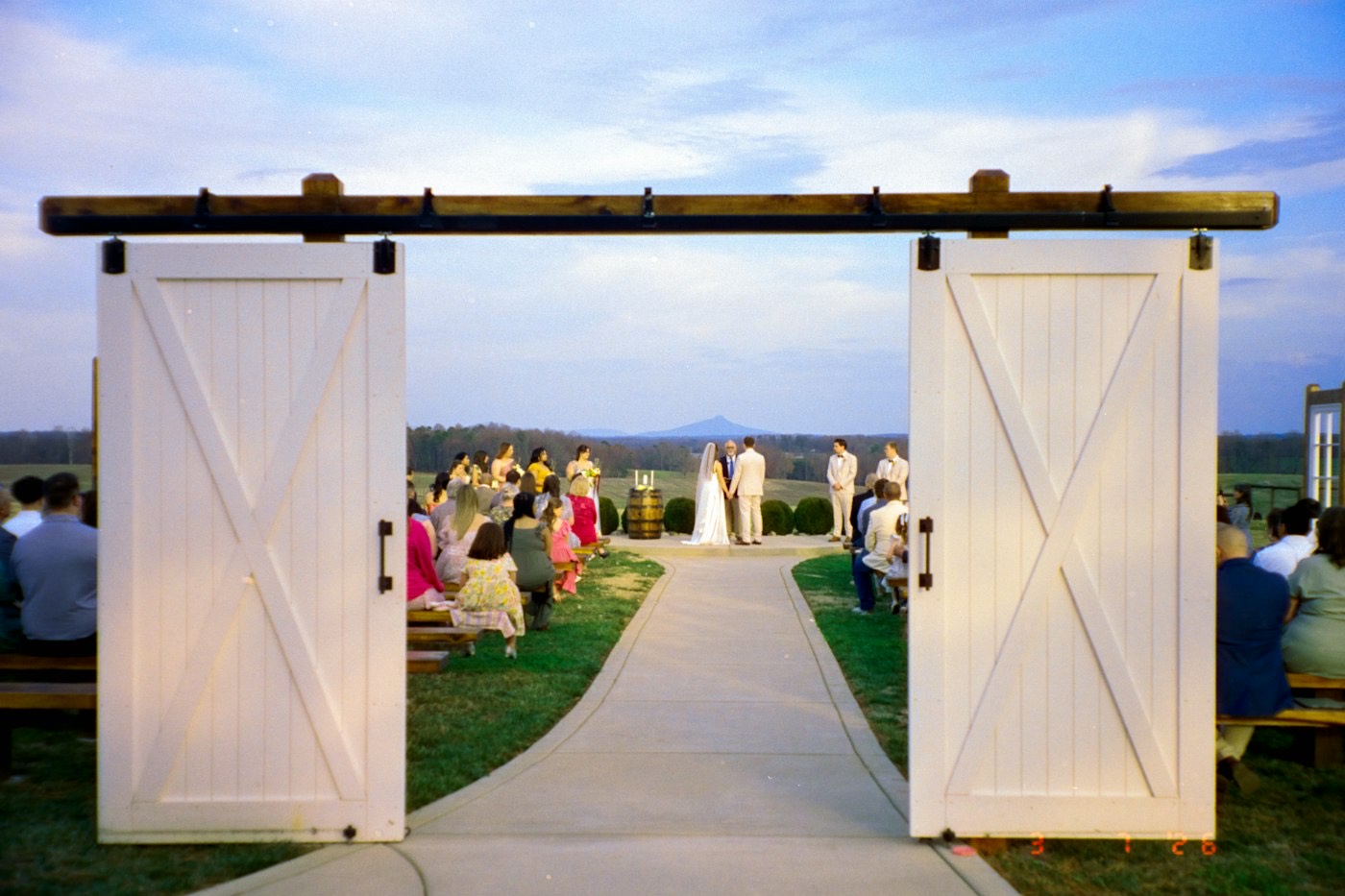 Open white barn doors frame an outdoor ceremony with guests on benches and a view of the hills